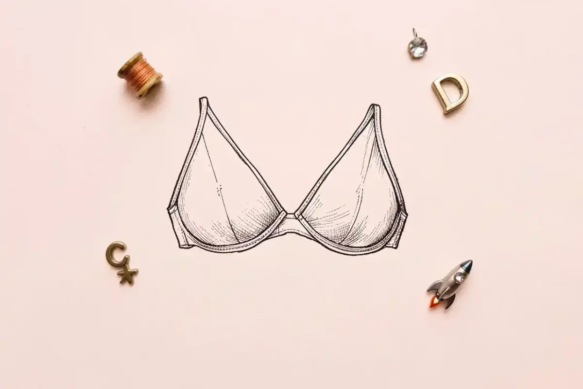 A vintage-style line drawing of a bra on a light pink background, surrounded by a spool of thread, letter charms, a rhinestone, and a tiny rocket