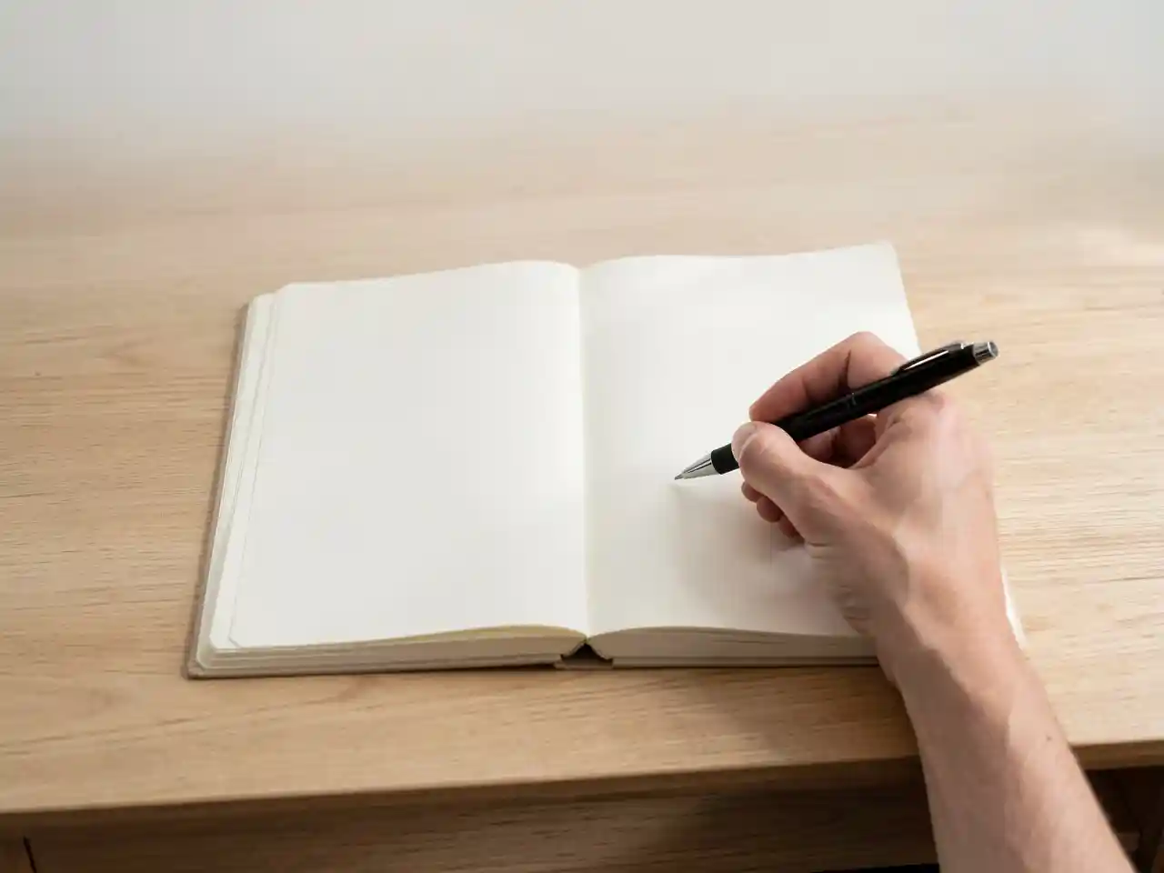 Hands holding a pen over a blank open notebook on a wooden desk in soft light