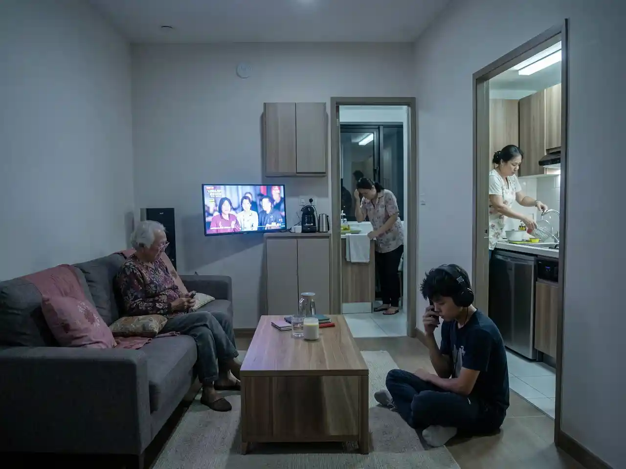 Three generations of a Filipino family in a cramped condo living room, each isolated by screens and tasks, symbolizing the emotional strain behind multigenerational living