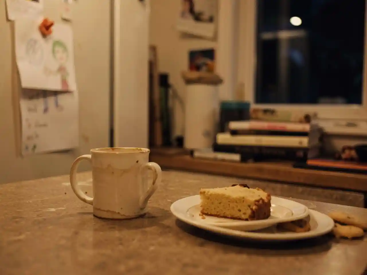 Warm, intimate kitchen countertop scene with a half-finished mug, a slice of cake, and a child's drawing on the fridge, symbolizing the kitchen as a witness to life's joys and sorrows