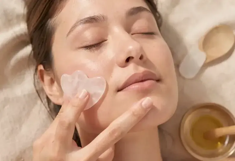 Close-up of a person's calm face with a hand gently gliding a rose quartz Gua Sha tool along the jawline, representing the science-backed practice of facial yoga and Gua Sha for non-invasive skin lifting