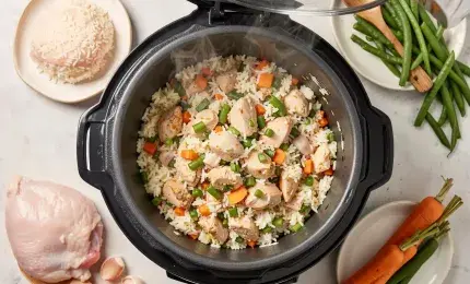 Overhead flat lay of an open rice cooker filled with chicken and vegetable rice, surrounded by fresh ingredients like raw chicken, rice, carrots, and beans