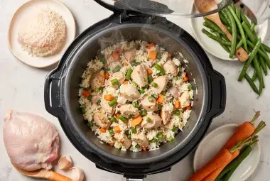 Overhead flat lay of an open rice cooker filled with chicken and vegetable rice, surrounded by fresh ingredients like raw chicken, rice, carrots, and beans