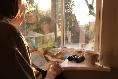 Person by a sunny window holding a map with binoculars and tea, looking out at the neighborhood