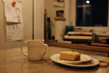 Warm, intimate kitchen countertop scene with a half-finished mug, a slice of cake, and a child's drawing on the fridge, symbolizing the kitchen as a witness to life's joys and sorrows